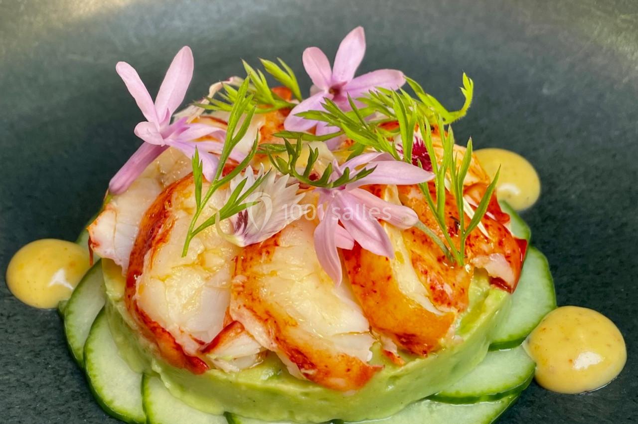 Plat raffiné composé de morceaux de homard, tranches de concombre, sauce jaune et fleurs comestibles en décoration.