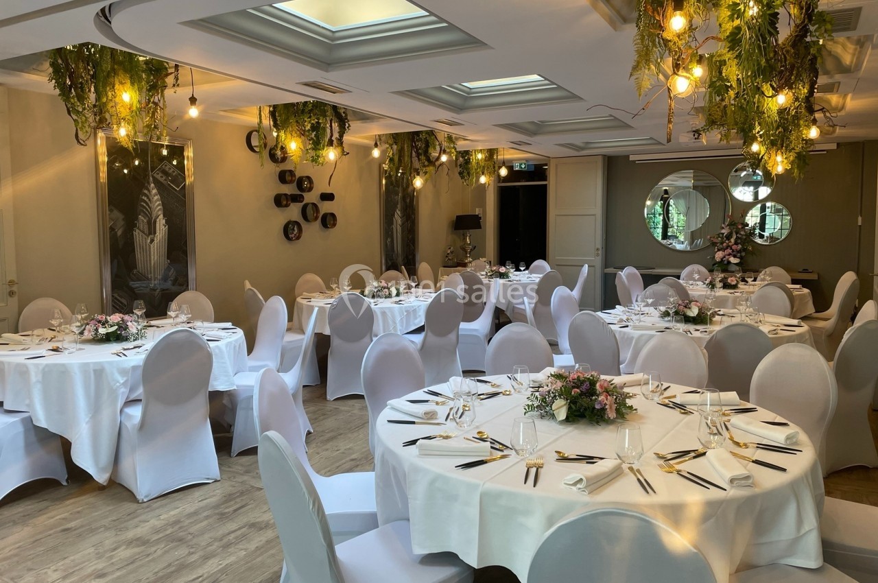 Salle de réception élégante avec tables rondes dressées, nappes blanches, décor floral et éclairage suspendu.
