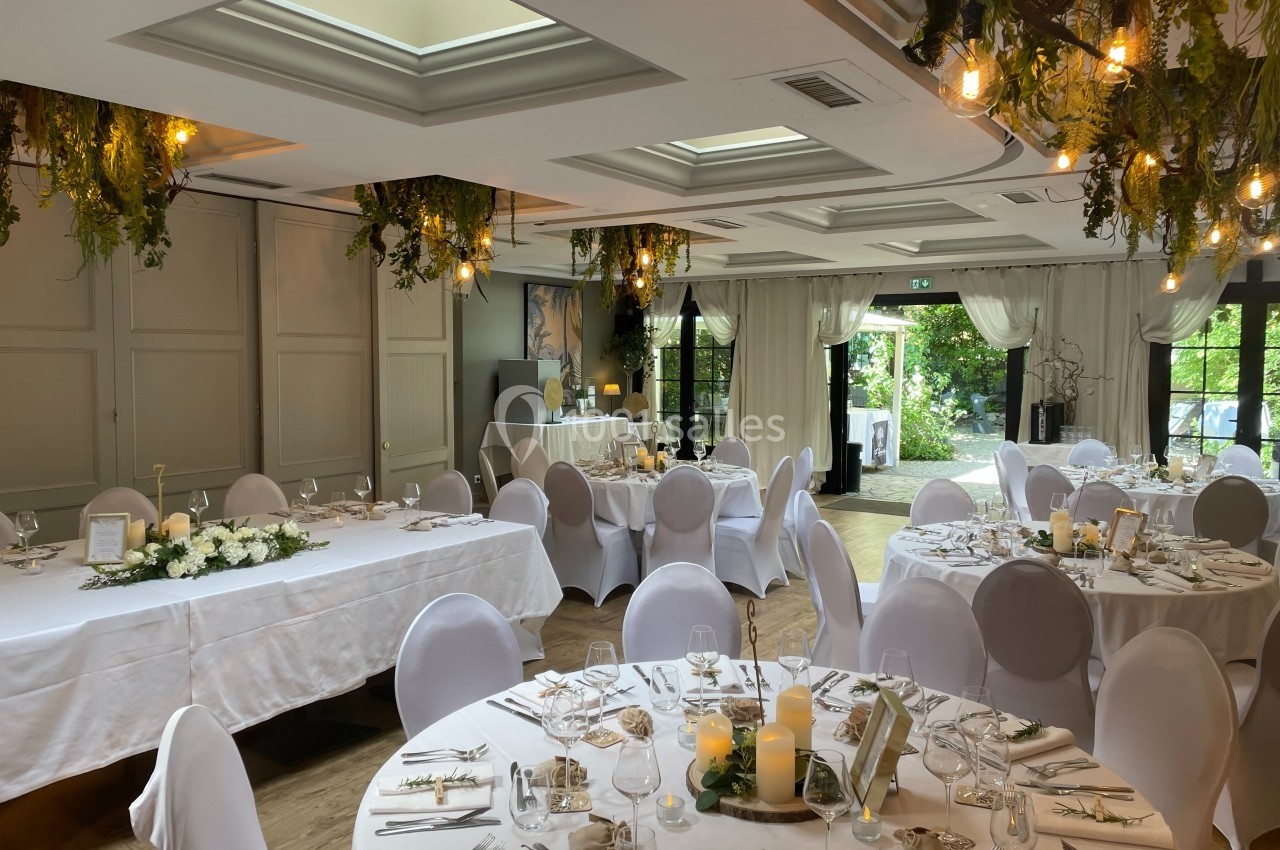 Salle de réception décorée pour un mariage, avec tables rondes dressées, guirlandes lumineuses et végétation suspendue.