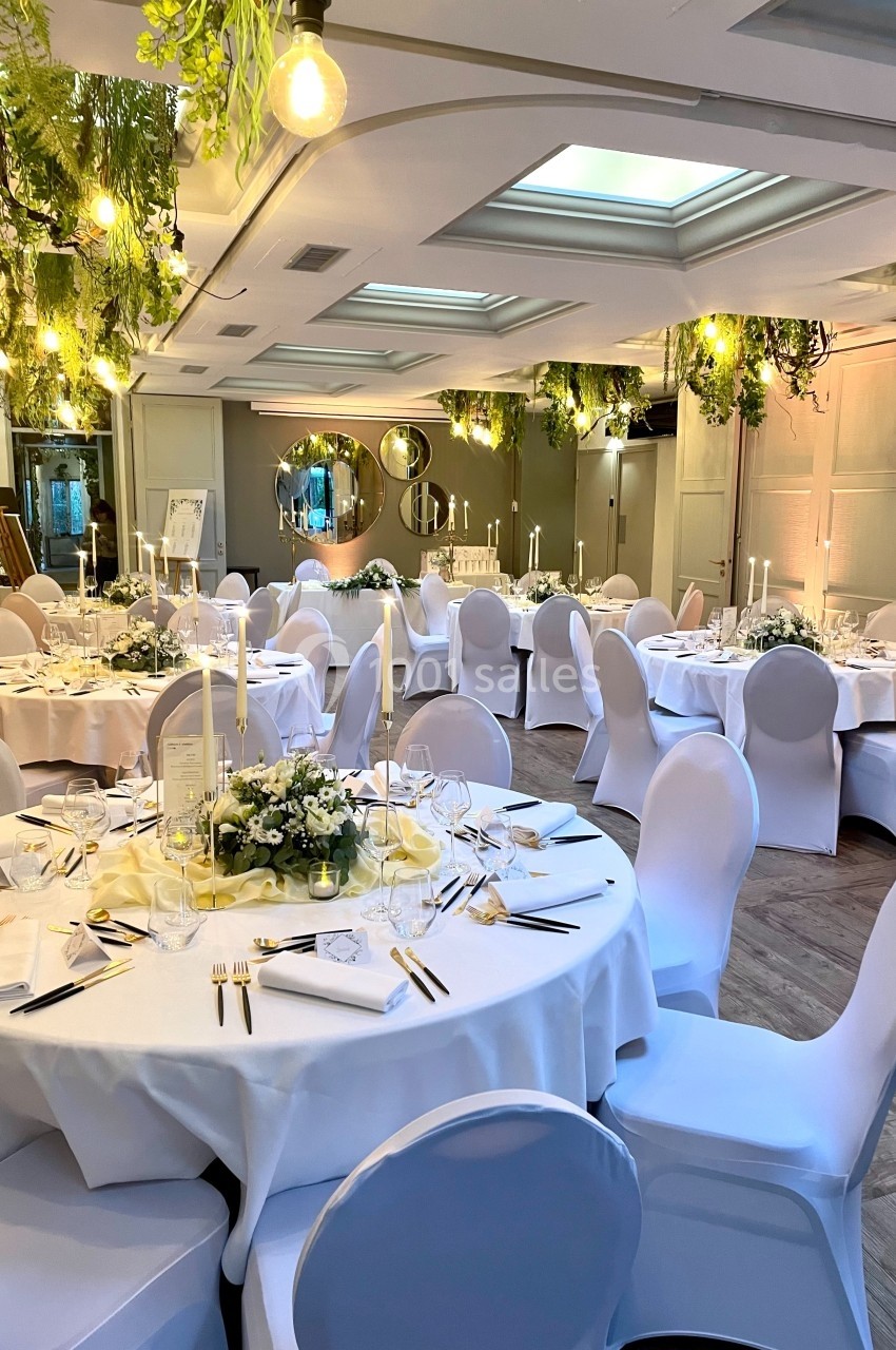 Salle de réception élégante avec tables rondes dressées, chaises blanches et décoration végétale suspendue.