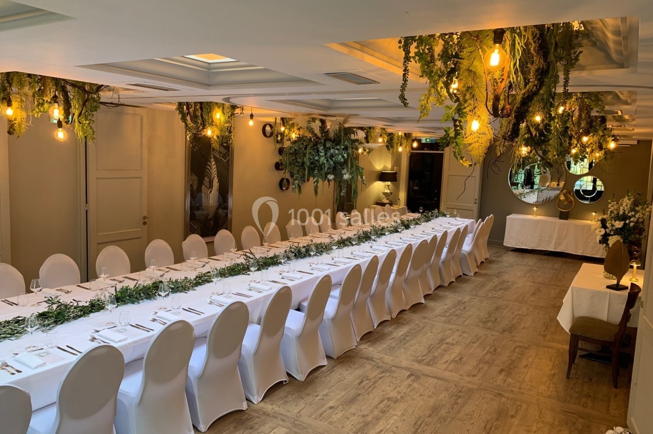 Salle de réception élégante avec longues tables dressées, chaises blanches et décoration végétale suspendue.