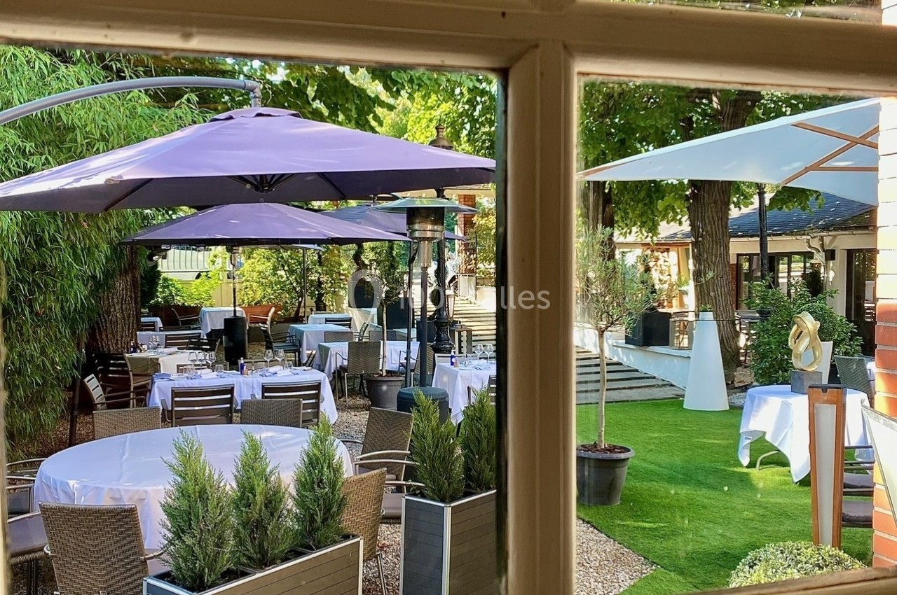 Terrasse extérieure avec tables dressées sous des parasols, entourée de verdure et visible à travers une fenêtre.