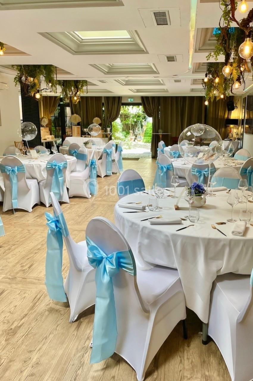 Salle de réception décorée avec des tables rondes, nappes blanches et chaises ornées de nœuds bleus.