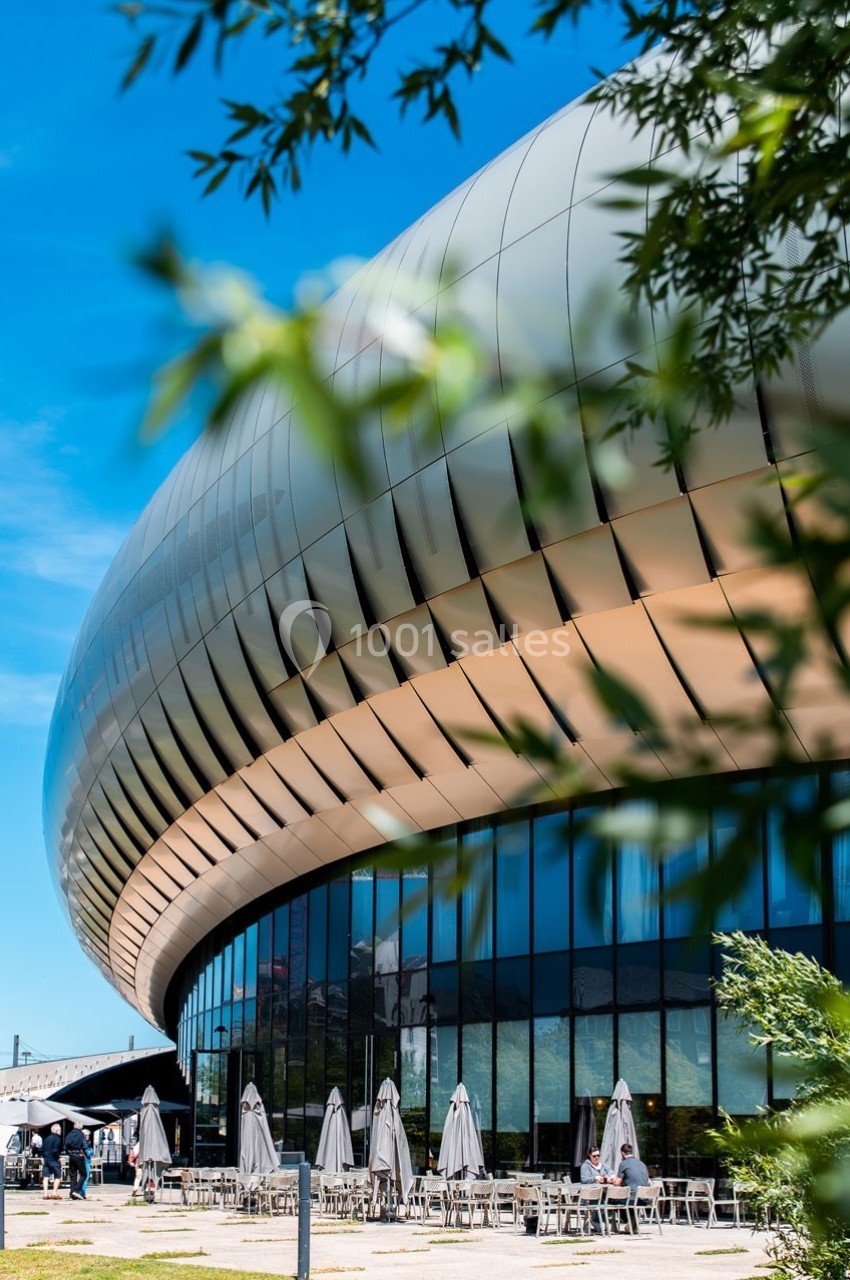 Façade moderne d'un bâtiment arrondi avec des baies vitrées, entouré de végétation et de parasols en terrasse.