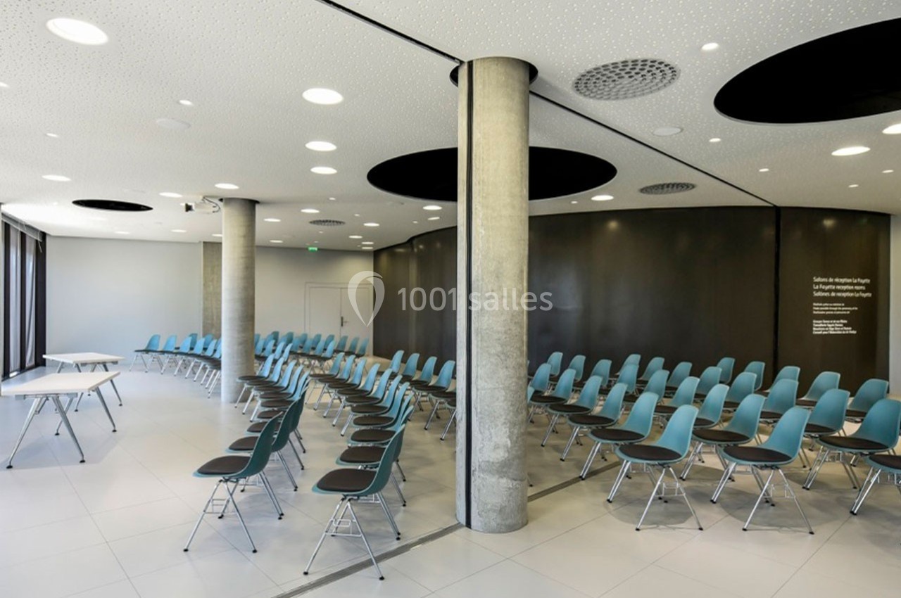 Salle de conférence moderne avec des rangées de chaises bleues, murs sombres et colonnes en béton apparent.