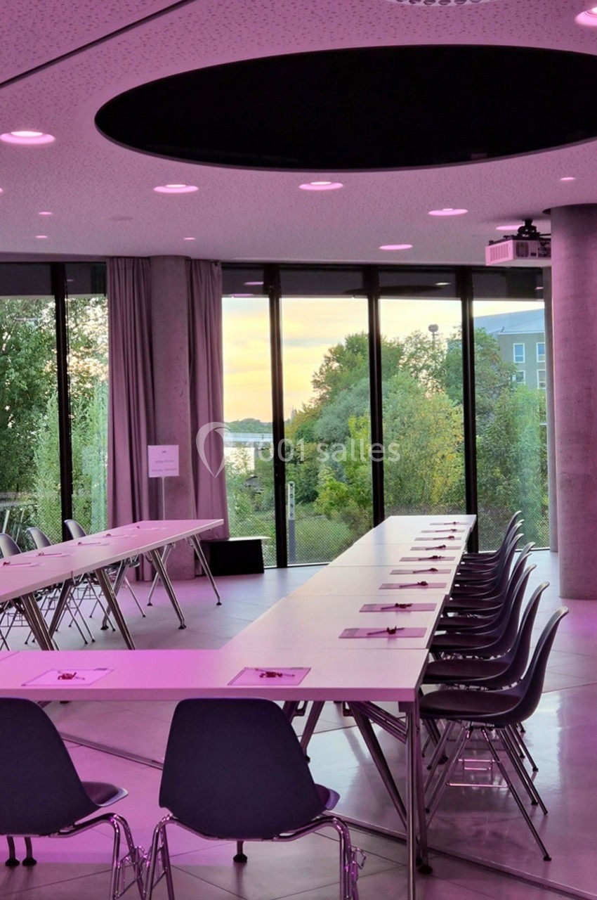 Salle de réunion moderne avec grandes baies vitrées, tables blanches alignées et chaises noires, vue sur un paysage…