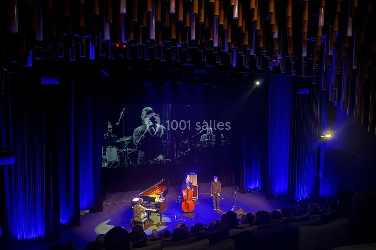 Un trio de musiciens joue sur scène dans une salle sombre, éclairée par des lumières bleues, avec un écran en arrière-plan.