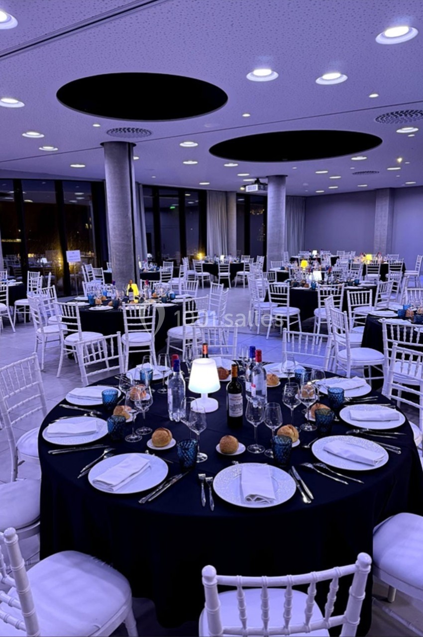 Salle de réception élégante avec tables rondes dressées, nappes noires, chaises blanches et éclairage tamisé.