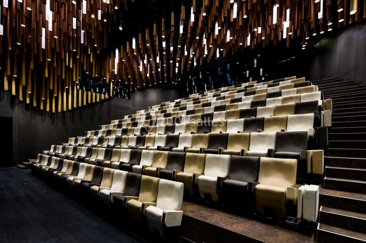 Salle de spectacle moderne avec des rangées de sièges beige et marron, éclairée par un plafond aux suspensions lumineuses.