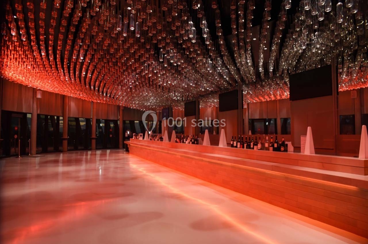 Plafond décoré de bouteilles suspendues, éclairage rouge, bar en bois long et moderne dans une grande salle.