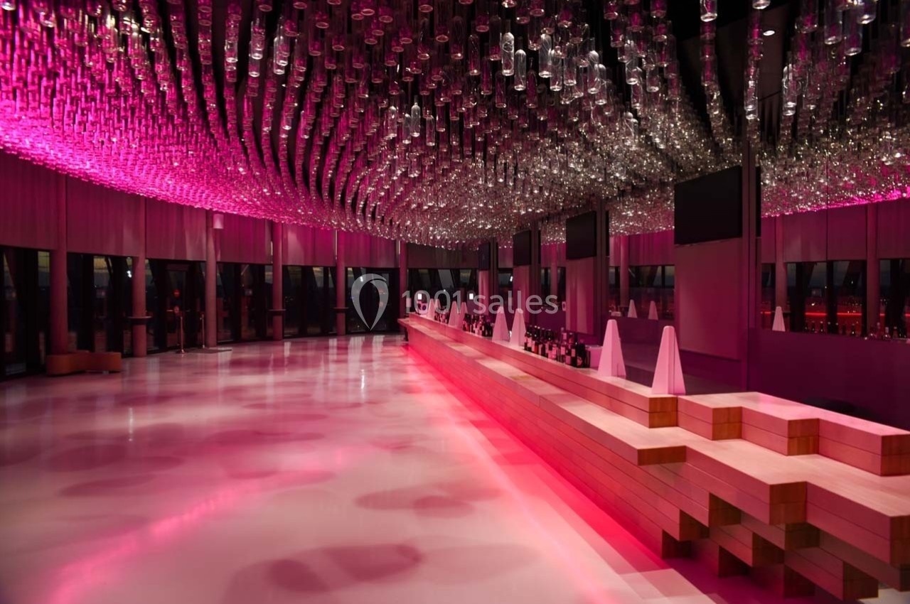 Salle moderne avec un long bar en bois, éclairée par des lumières roses et un plafond orné de suspensions en verre.