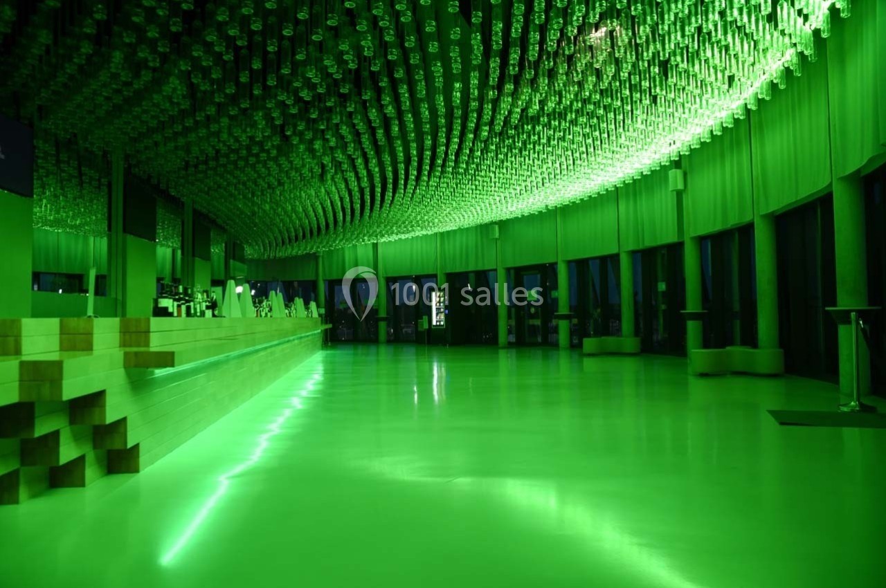 Salle moderne éclairée par une lumière verte, avec un bar à gauche et un plafond décoré de suspensions cylindriques.