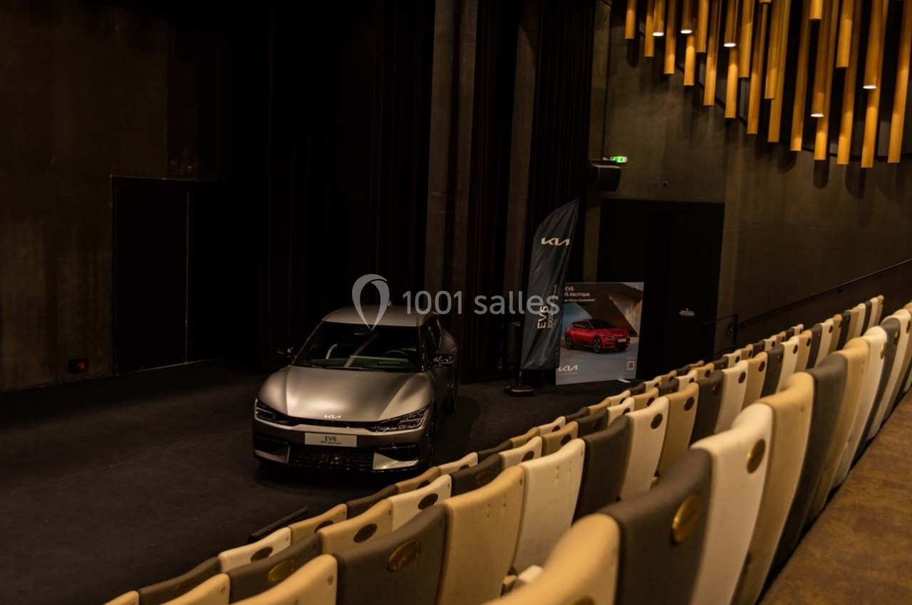 Une voiture grise exposée sur une scène dans une salle sombre avec des rangées de sièges et un panneau promotionnel.