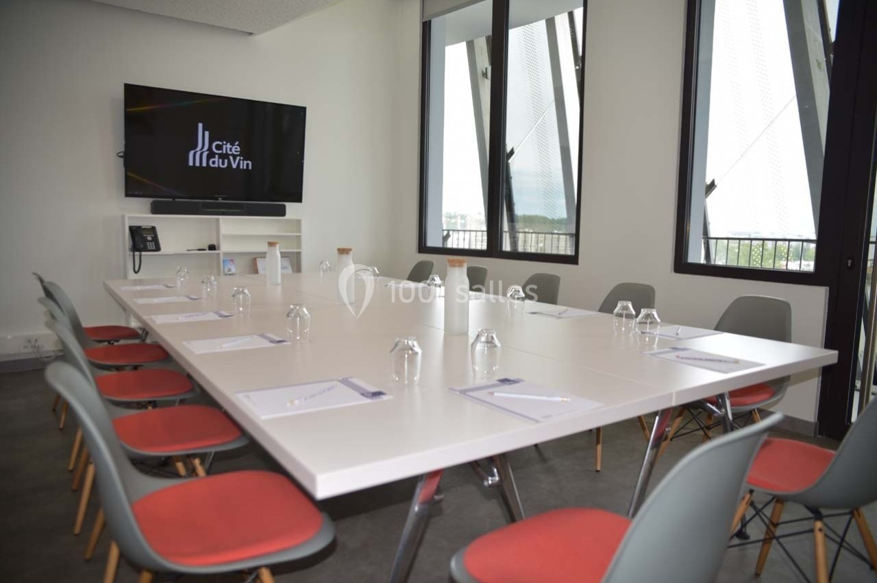 Salle de réunion lumineuse avec une grande table, des chaises rouges, un écran mural et des fenêtres donnant sur l'extérieur.