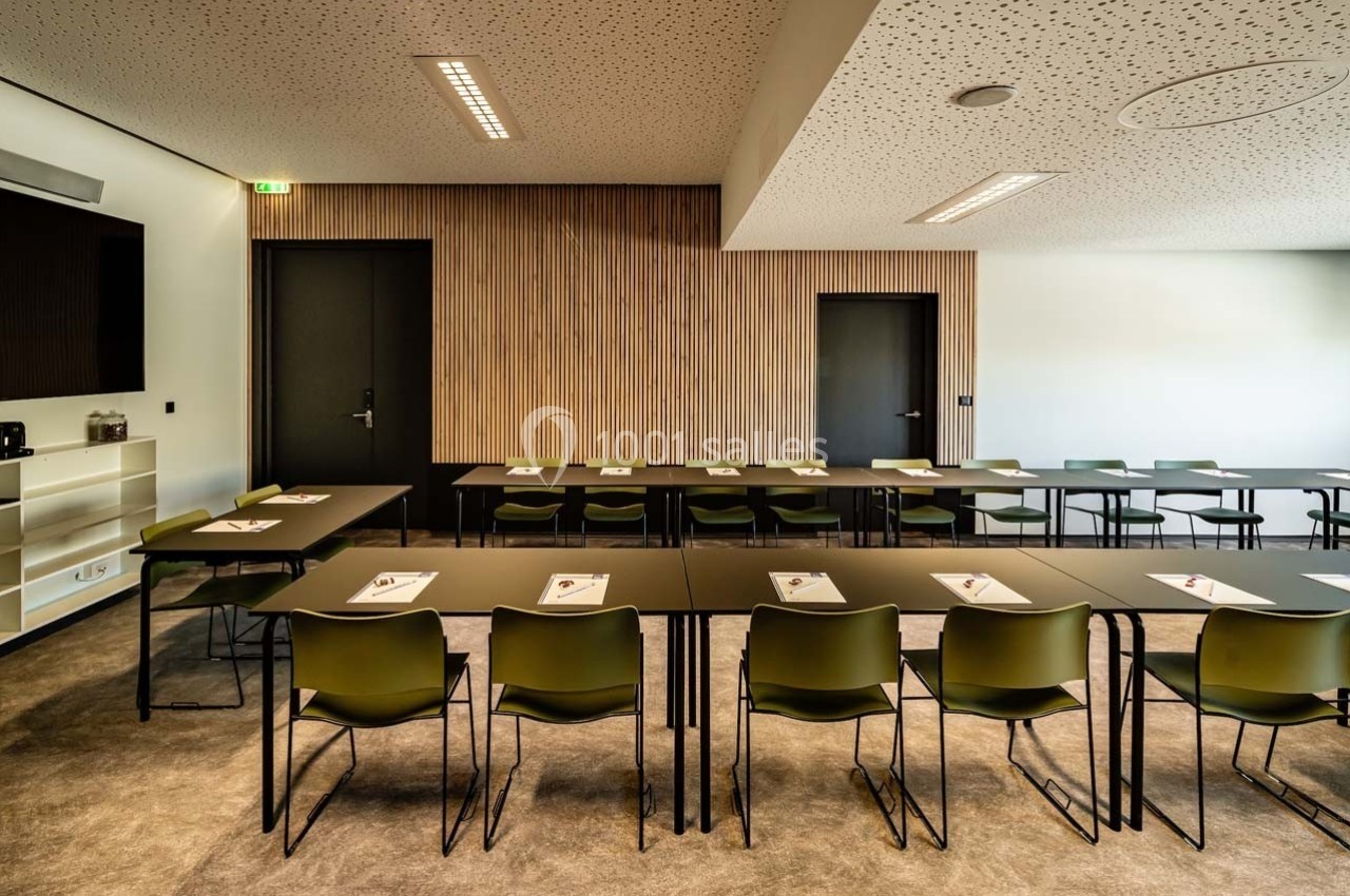 Salle de réunion moderne avec tables noires, chaises vertes et documents posés, éclairée par des plafonniers.