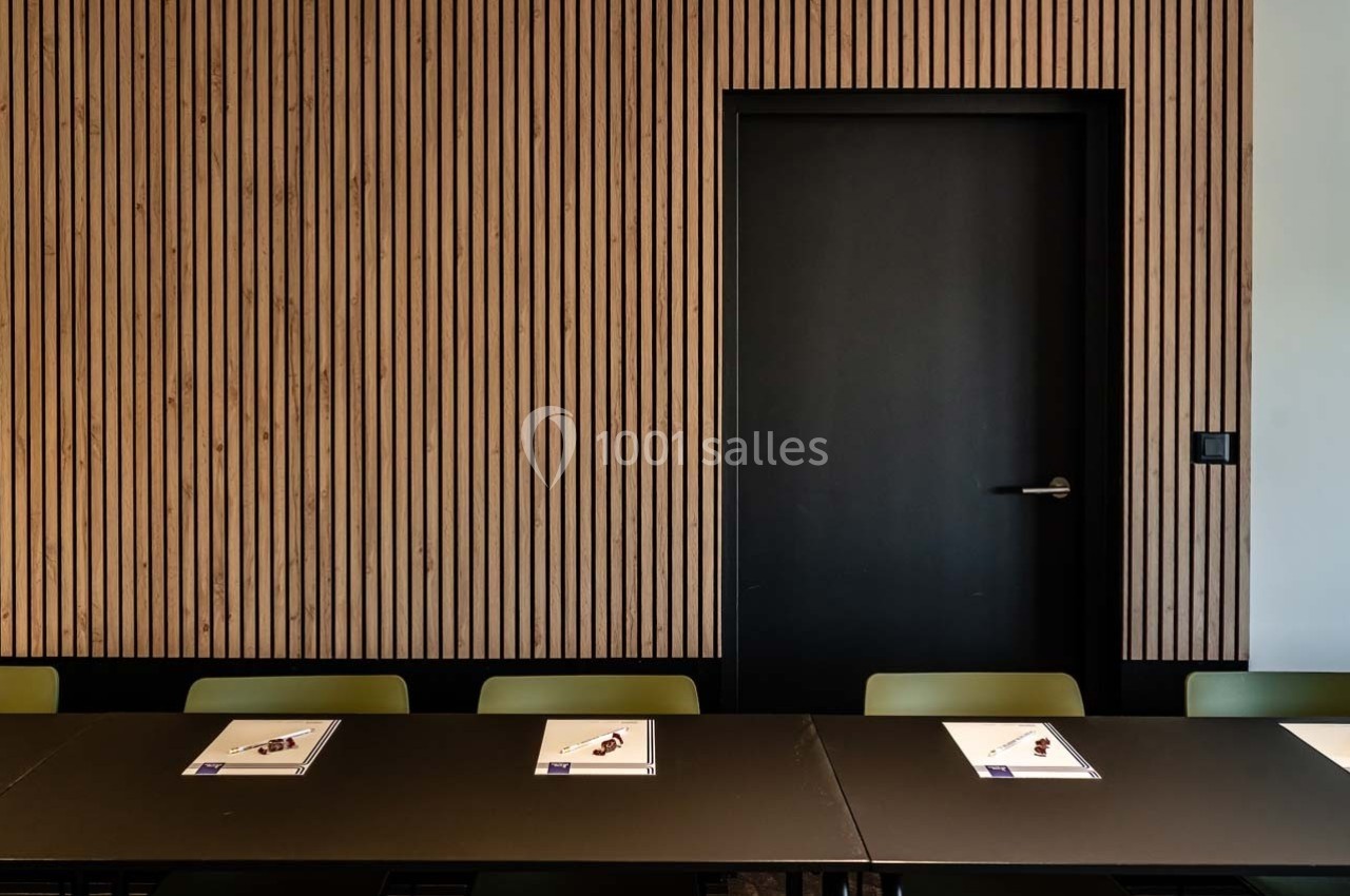 Salle de réunion avec tables noires, chaises vertes et mur en bois à lattes, porte noire fermée en arrière-plan.