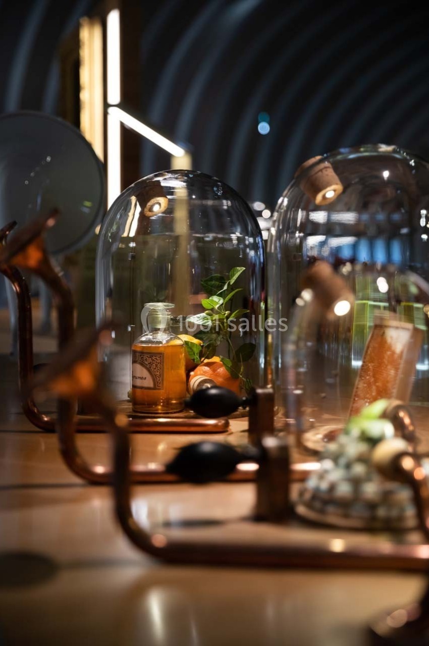 Flacons et éléments décoratifs sous cloches en verre sur une table éclairée dans un espace moderne et tamisé.