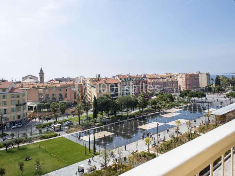 Vue d'une place avec fontaines, espaces verts et bâtiments colorés, prise depuis un balcon en hauteur.