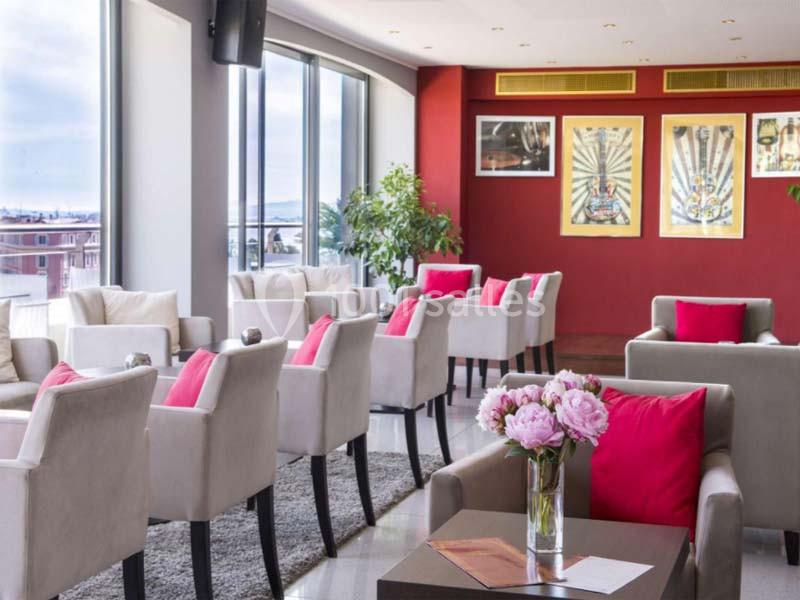 Salle lumineuse avec fauteuils beiges, coussins rouges, tables basses, grandes fenêtres et décorations murales colorées.