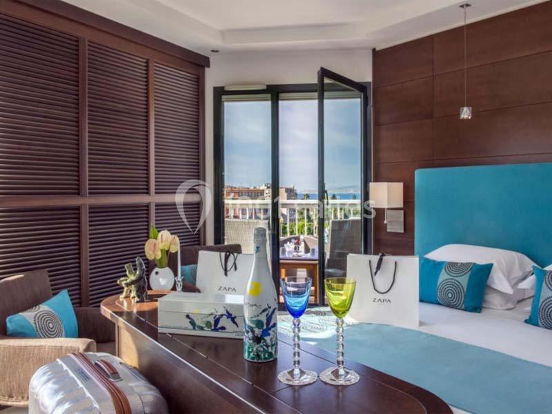 Chambre d'hôtel lumineuse avec lit double, balcon ouvert sur une vue urbaine, et décorations modernes sur une table.