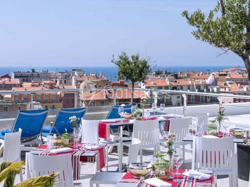 Terrasse d'un restaurant avec tables dressées, vue sur des toits de ville et la mer en arrière-plan.