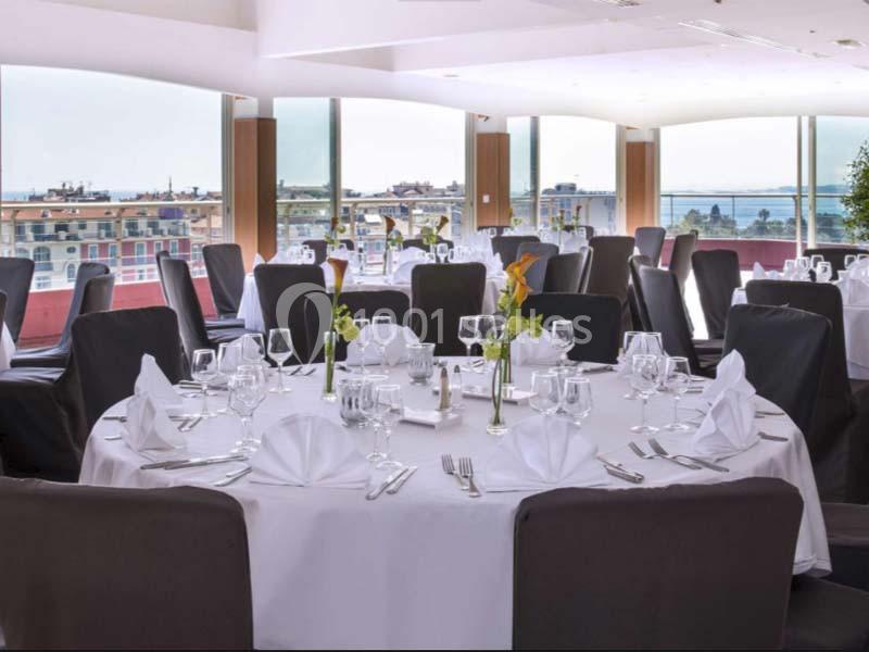 Salle de réception lumineuse avec tables rondes dressées, nappes blanches et vue sur la ville et la mer.