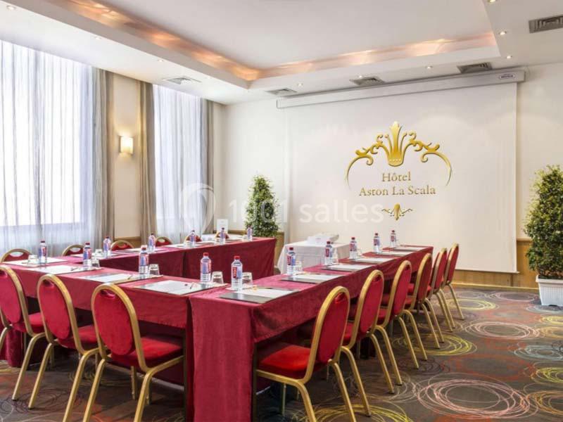Salle de réunion lumineuse avec tables disposées en U, chaises rouges et décorations élégantes.