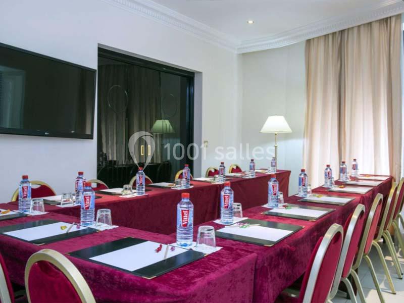 Salle de réunion équipée de tables recouvertes de nappes rouges, chaises alignées, bouteilles d'eau et blocs-notes.