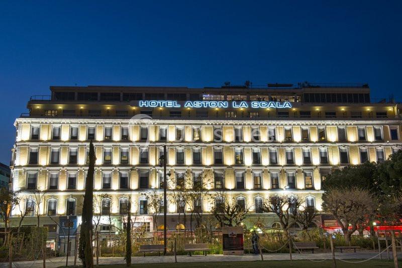 Façade illuminée de l'hôtel Aston La Scala de nuit, avec des arbres et un espace vert au premier plan.