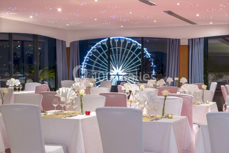 Salle de réception élégante avec tables dressées, vue sur une grande roue illuminée à travers des baies vitrées.