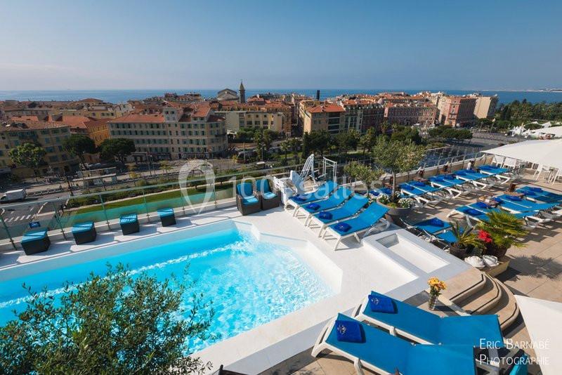 Vue d'une piscine extérieure sur une terrasse surplombant une ville côtière avec des bâtiments et la mer en arrière-plan.