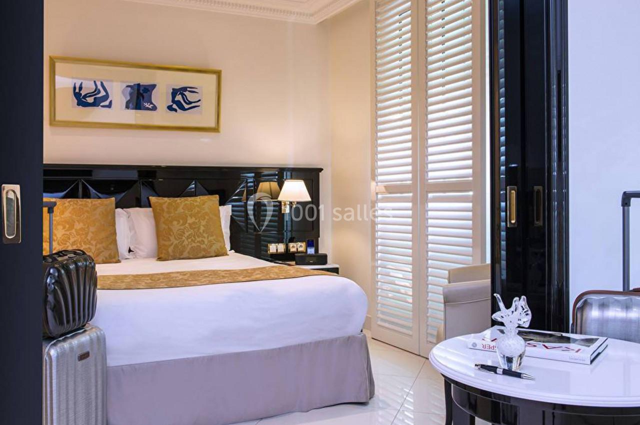 Chambre d'hôtel lumineuse avec lit double, coussins dorés, valises, table basse et volets blancs.