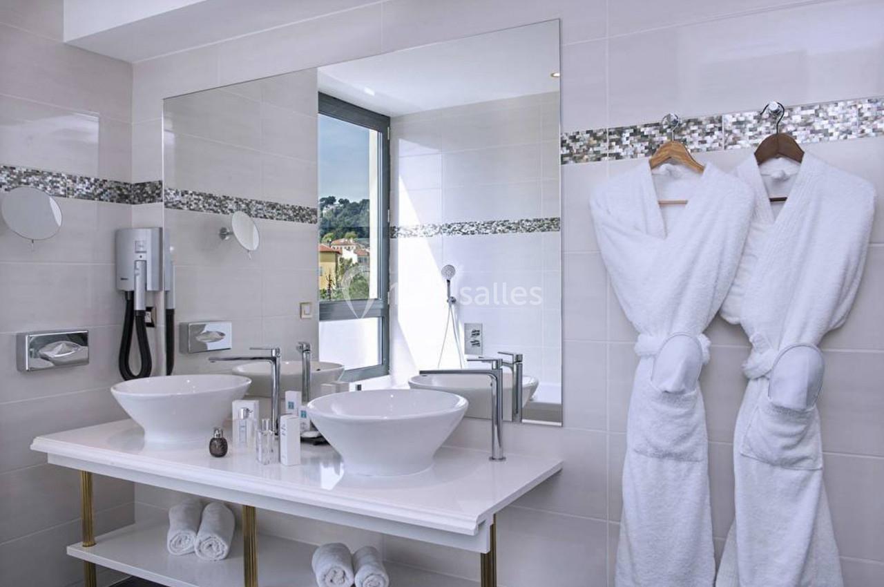 Salle de bain moderne avec deux vasques, miroirs, peignoirs suspendus, et fenêtre offrant une vue extérieure.