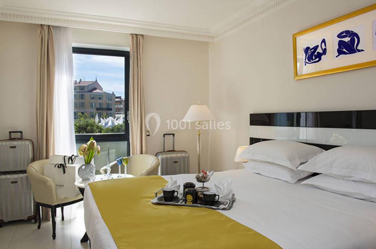 Chambre d'hôtel lumineuse avec lit double, table basse, valises, et vue sur des bâtiments par une fenêtre.