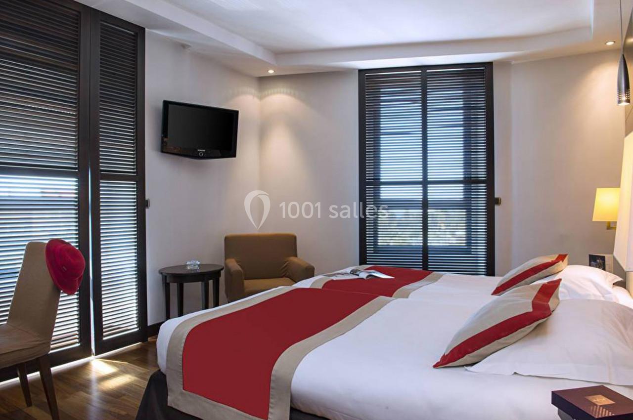 Chambre d'hôtel lumineuse avec deux lits simples, fauteuil, table, téléviseur mural et grandes fenêtres avec stores.