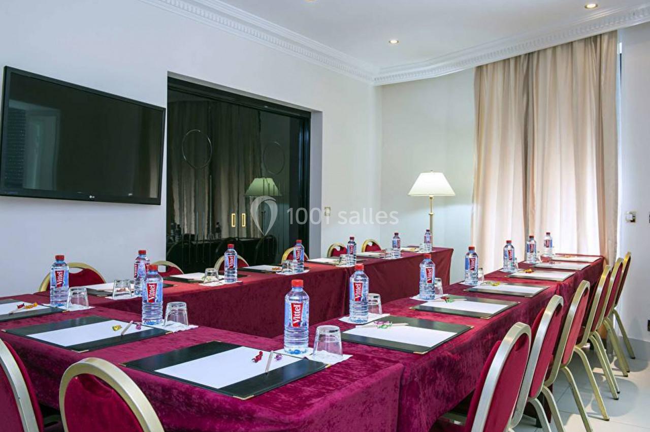 Salle de réunion équipée de tables en U avec nappes rouges, bouteilles d'eau, blocs-notes et écran mural.