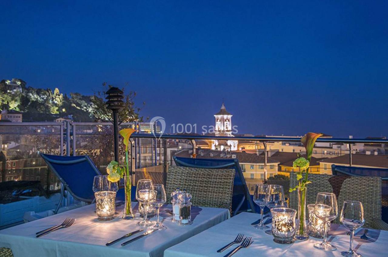 Tables dressées avec chandelles et fleurs sur une terrasse offrant une vue nocturne sur des bâtiments éclairés.