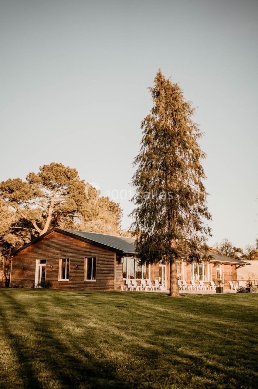 Maison en bois entourée d'un jardin verdoyant avec un grand arbre au premier plan, sous un ciel clair.