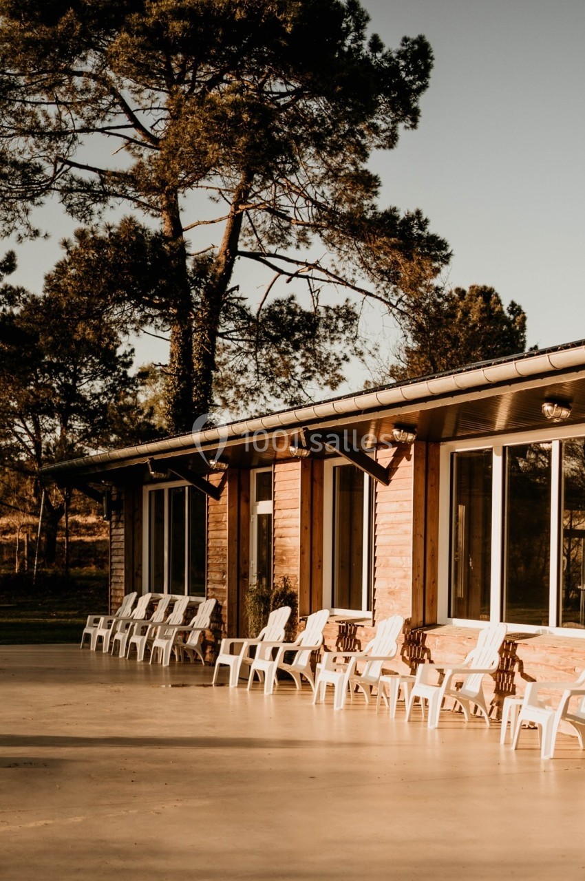 Façade d'un bâtiment en bois avec de grandes fenêtres, entouré de chaises blanches et d'arbres sous une lumière douce.