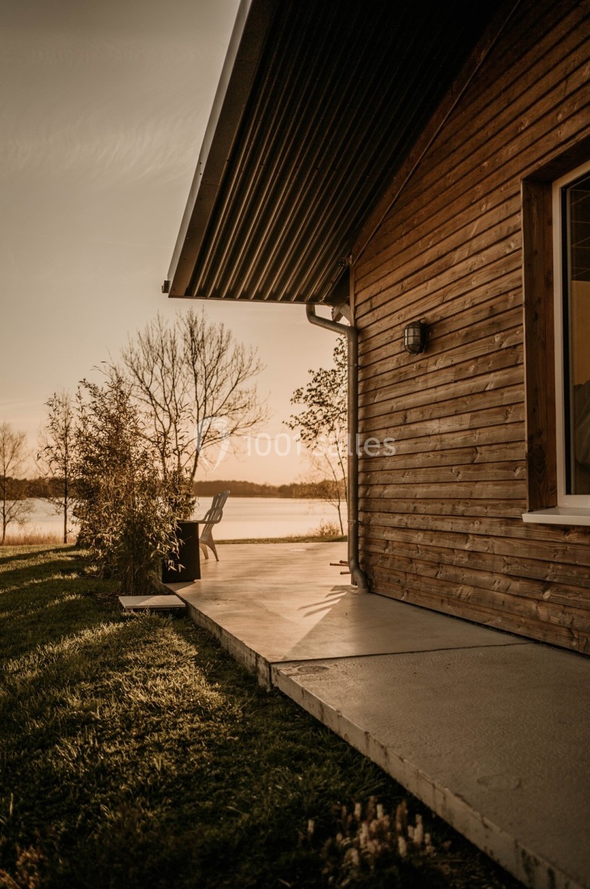 Façade en bois d'une maison avec terrasse, vue sur un lac au coucher du soleil et arbres en arrière-plan.