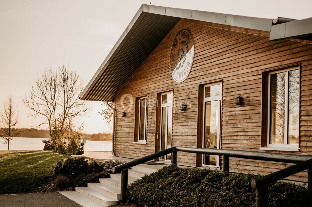 Bâtiment en bois avec de grandes fenêtres, situé près d'un plan d'eau et entouré de végétation.