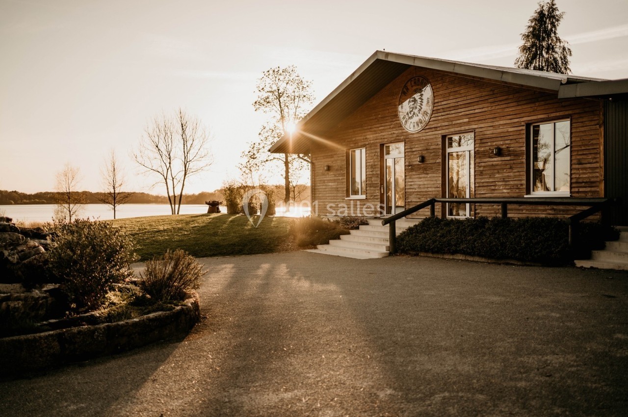 Maison en bois avec grande baie vitrée, entourée de végétation, au bord d'un lac au coucher du soleil.
