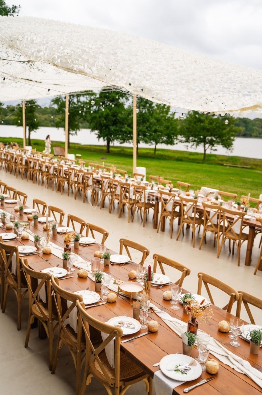 Tables en bois décorées pour un repas en plein air sous une toile blanche, avec vue sur un lac et des arbres.