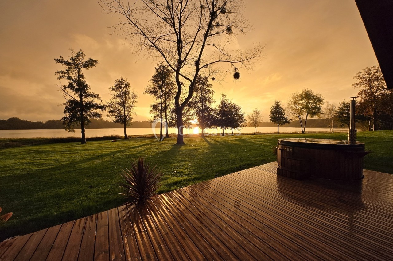 Coucher de soleil sur un lac, avec des arbres en contre-jour et une terrasse en bois au premier plan.