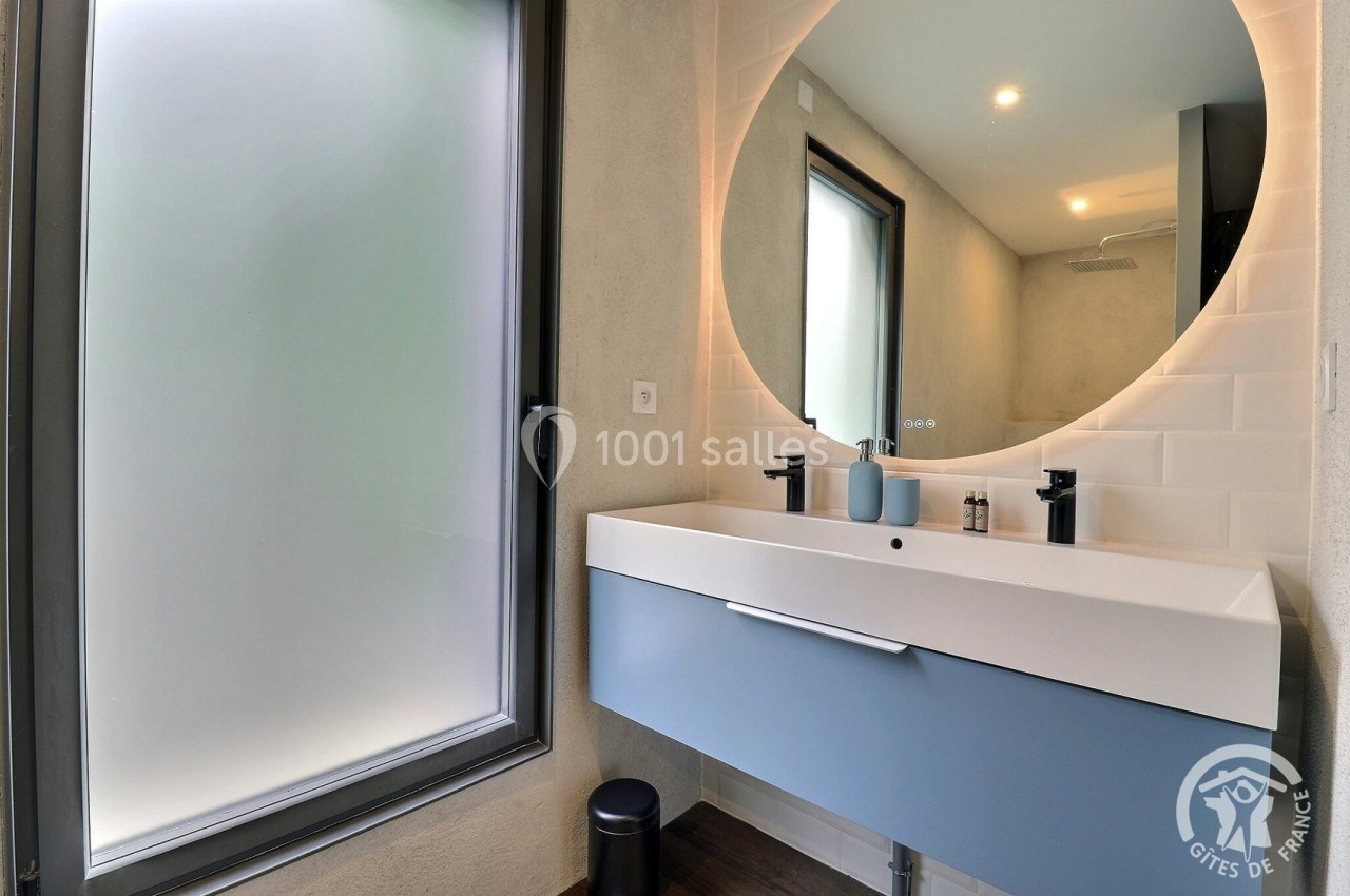 Salle de bain moderne avec lavabo suspendu, miroir rond éclairé, fenêtre givrée et douche en arrière-plan.