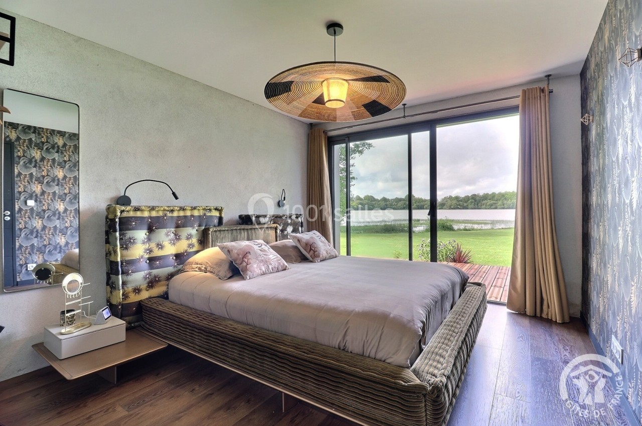 Chambre moderne avec lit double, grande baie vitrée donnant sur un lac et décoration élégante.