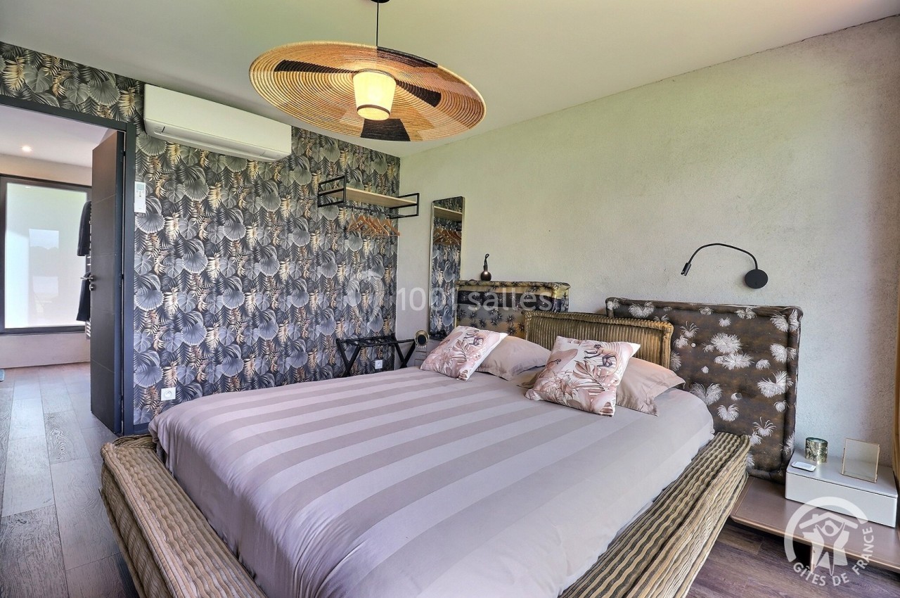 Chambre avec lit double, décoration murale tropicale, suspension en osier et lumière naturelle provenant d'une porte vitrée.