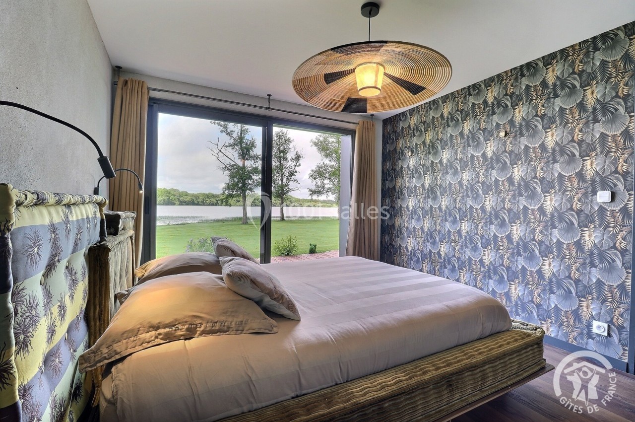 Chambre avec lit double, grande baie vitrée donnant sur un paysage de lac et mur décoré de papier peint à motifs.