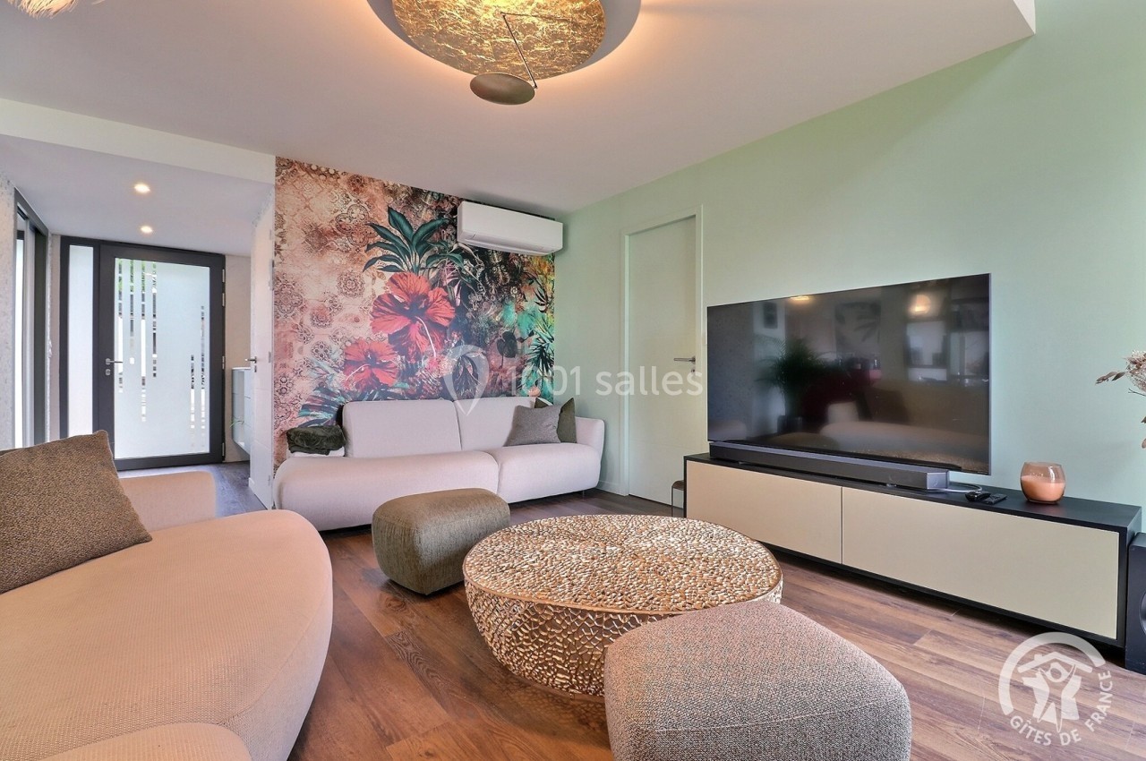Salon moderne avec canapé beige, table basse ronde, mur décoré de motifs floraux et télévision sur meuble clair.