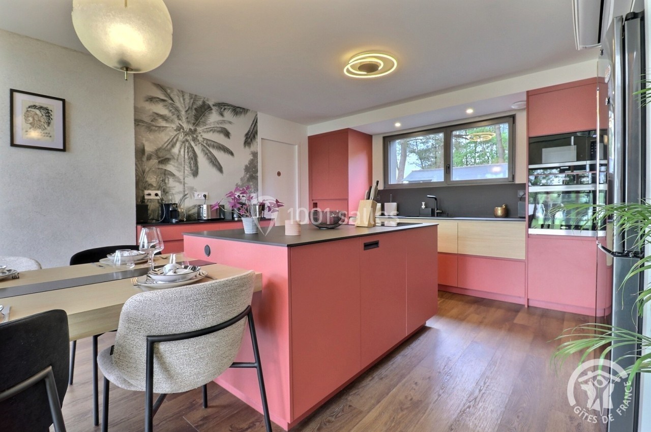 Cuisine moderne avec îlot central rose, table dressée pour un repas, parquet en bois et grande fenêtre apportant de la…